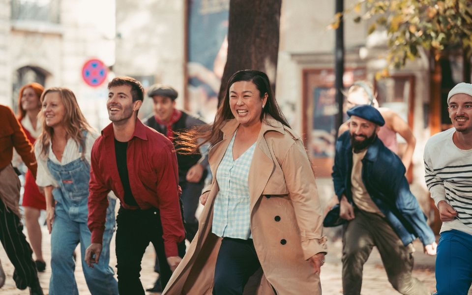 A group of people smile and dance together in a sun-lit town square.
