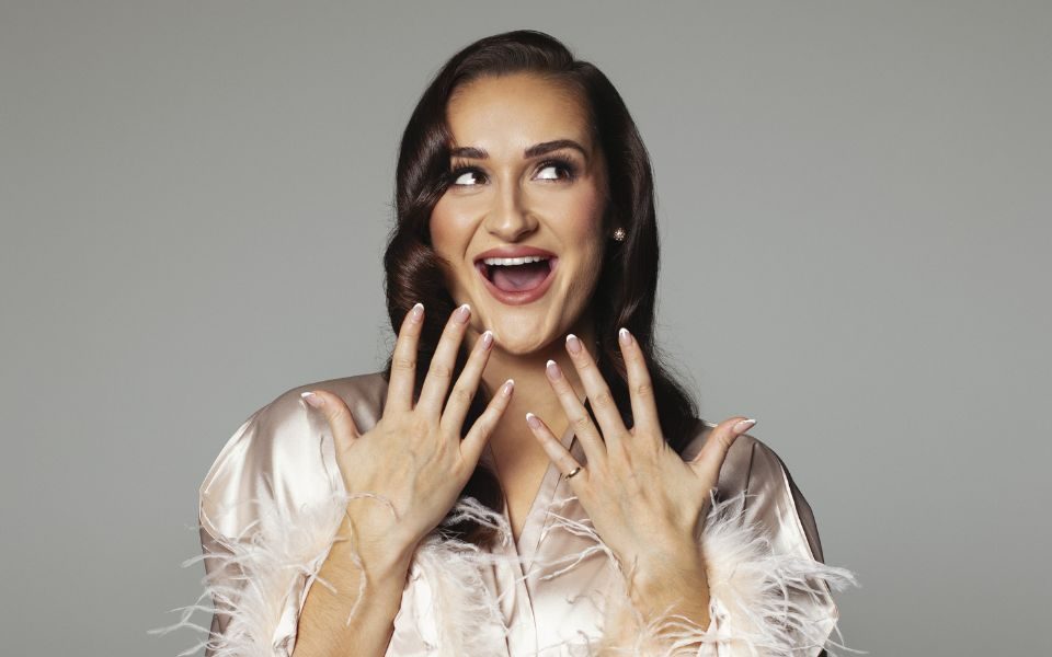 Maddie Grace Jepson smiles with an open mouth and looks off camera, holding up manicured hands. She wears a silk dressing gown with feathery sleeves and makeup.