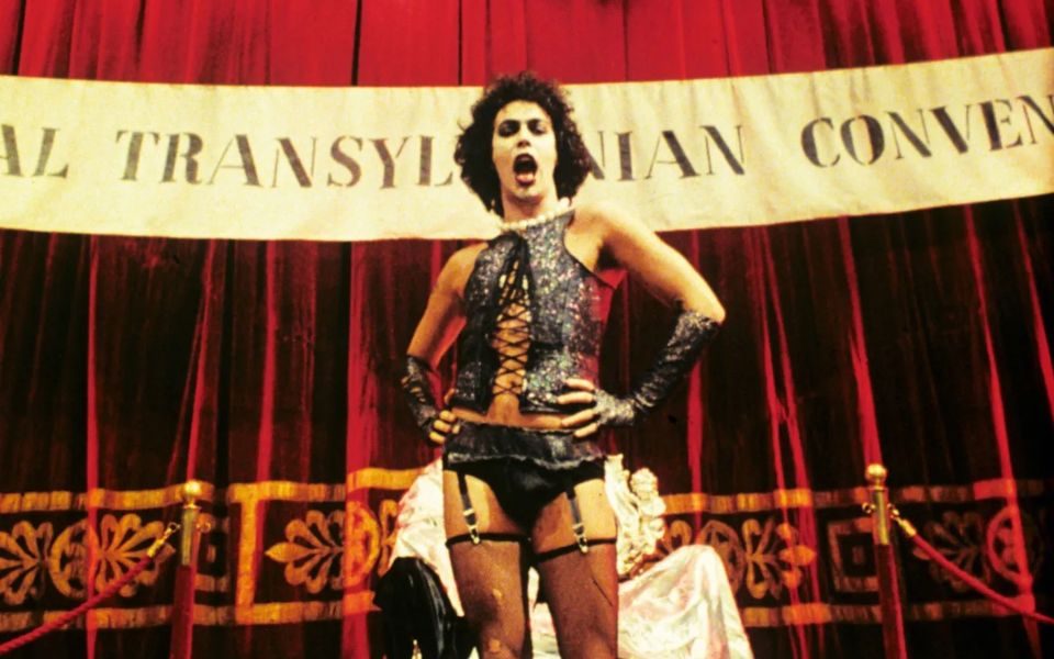 Tim Curry as Dr. Frank-N-Furter confidently sings with hands on hips in front of a red curtain, wearing glittery lingerie and fishnets.