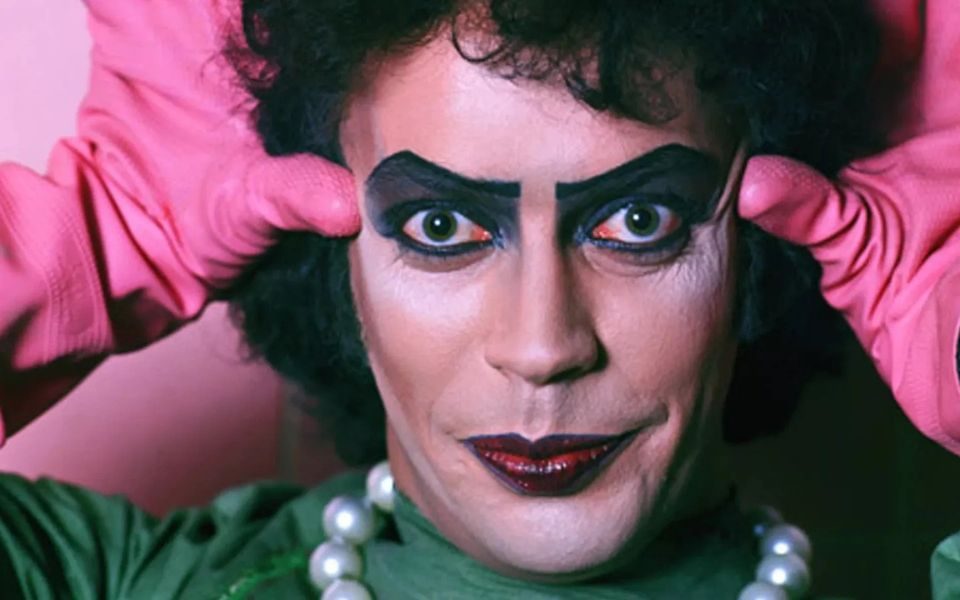 Tim Curry from The Rocky Horror Picture Show in close-up wearing heavy, stylised makeup and bead necklace, holds his pink gloved hands up to his head.