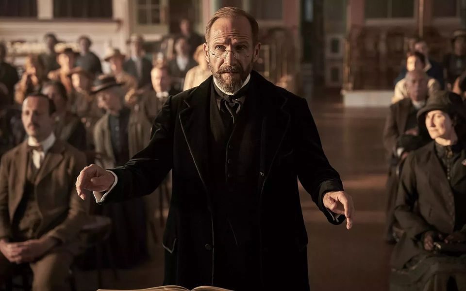 In early twentieth century clothing and spectacles, Ralph Fiennes speaks in front of a crowd of people sat together in a hall.