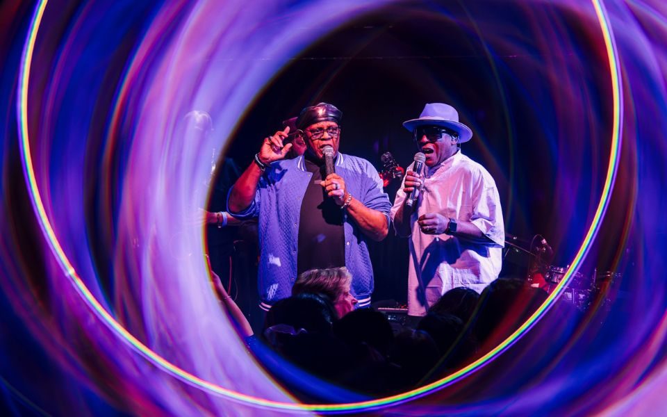 Two singers with microphones perform on stage, surrounded by a soft halo of purple light.