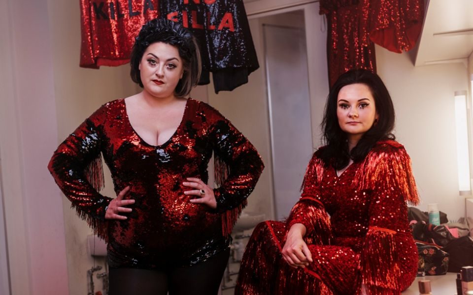 In red and black sequined outfits, the two members of All Killa No Filla sassily face the camera in a dressing room, which features two sequined outfits in the background reading 'All Killa' and 'No Filla'.