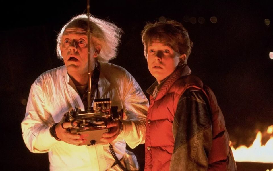 White-haired scientist Doc Brown (Christopher Lloyd) holds onto a sci-fi remote control with a look of anticipation, alongside Michael J Fox as Marty McFly.
