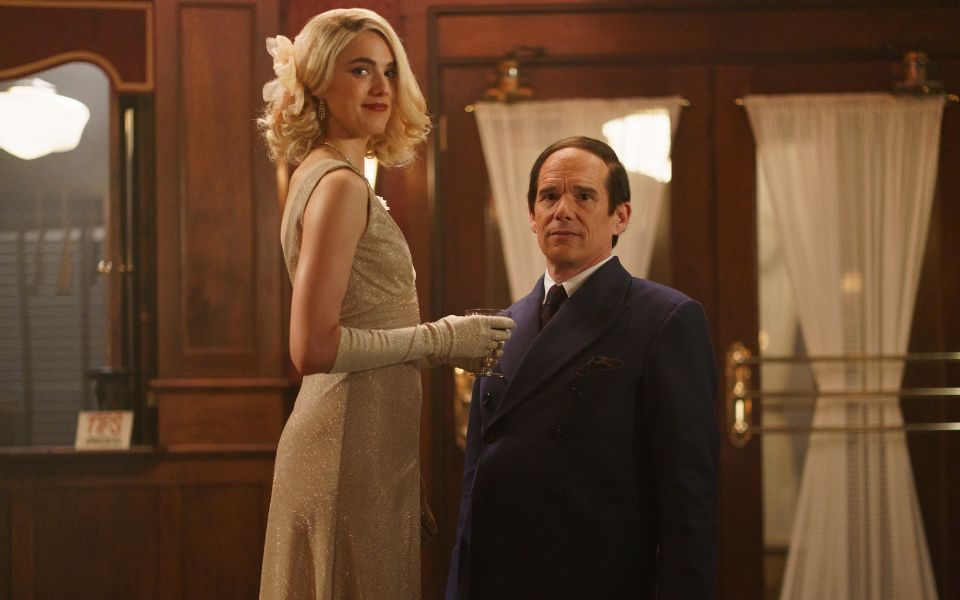 Ethan Hawke with combed hair in a vintage suit alongside a blonde-haired Margaret Qualley in long gloves and a glittery dress, holding an elegant glass.