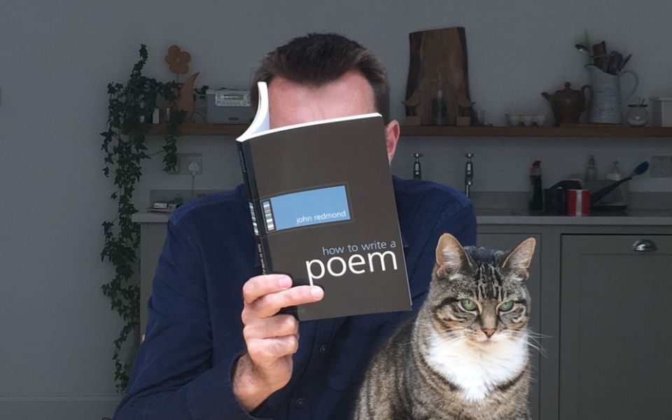 At a kitchen counter beside a tabby cat, Brian Bilston reads a book entitled 'how to write a poem', his face obscured behind its cover.