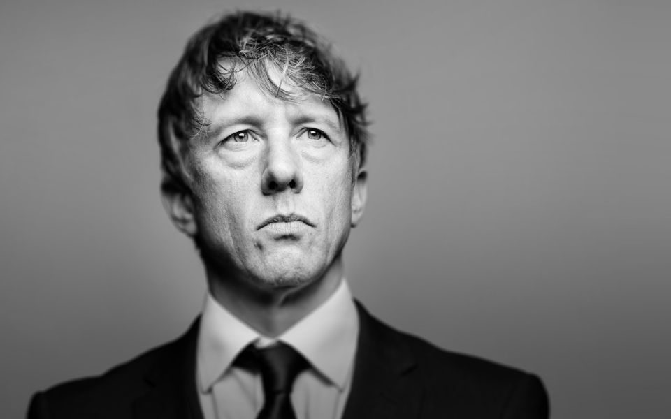 In black and white, Jonathan Pie looks sternly off-camera, standing poised in a black suit.