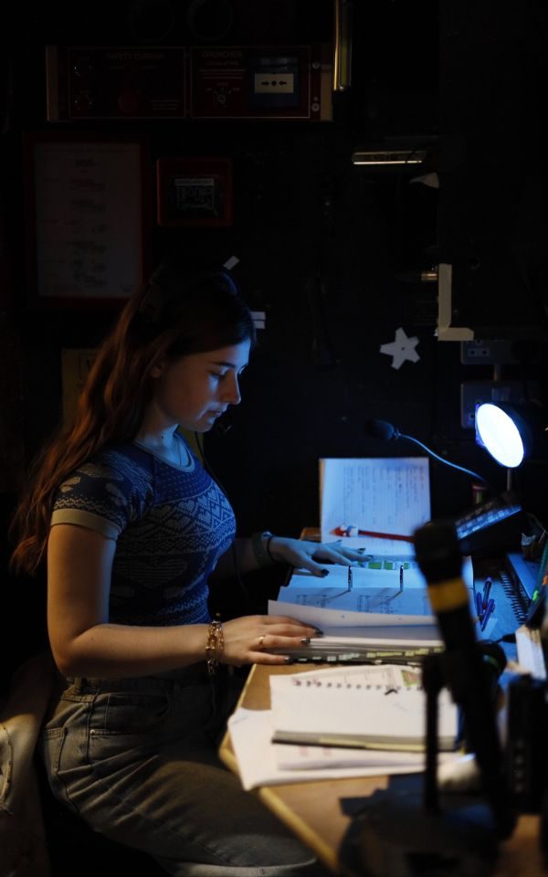 Joni Franieczek sat at a desk reading a score in a ringbinder while surrounded by notes and a mic for backstage comms. She is lit by a desklamp in the dimly lit backstage area.