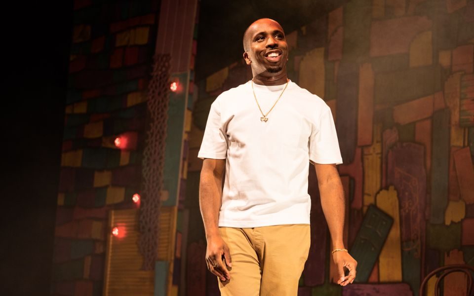 Kiell Smith Bynoe smiles onstage, wearing a white t-shirt and gold pendant chain under warm lighting.