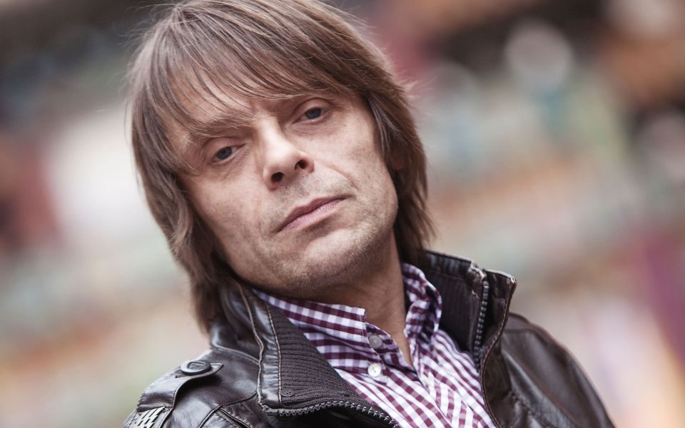 At a tilted Dutch angle, Mani stands in a leather jacket and shirt in front of a blurred background.