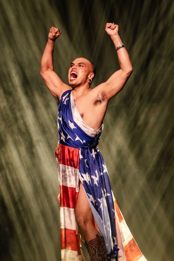 Seann Miley Moore as the Engineer wearing an American-flag print dress raising his fists in the air as he sings.