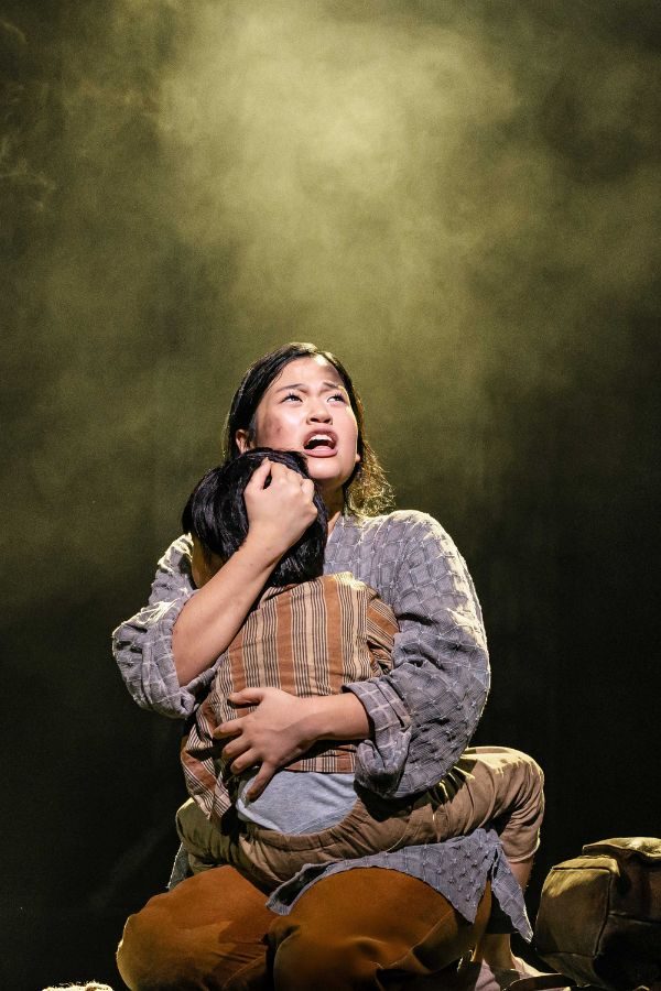 A woman in battered grey and brown clothing kneels on the floor clutching her small child in her arms as she sings.