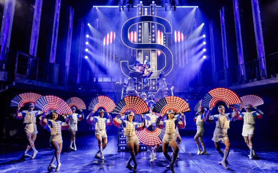 A chorus of ensemble members hold up giant fans with American flags on them. Above them, The Engineer character sits on a giant dollar sign in an American flag costume.