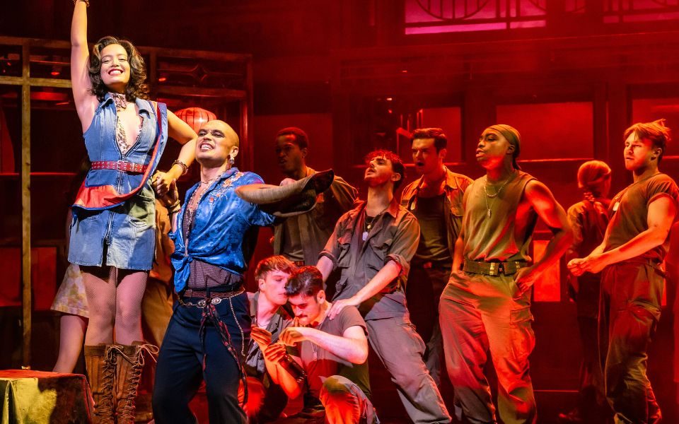Ace as Gigi in a denim mini-dress and brown boots standing proudly on a stool holding hands with Seann Miley Moore as the Engineer. Behind them a group of American GIs stare at her in their khaki uniforms.