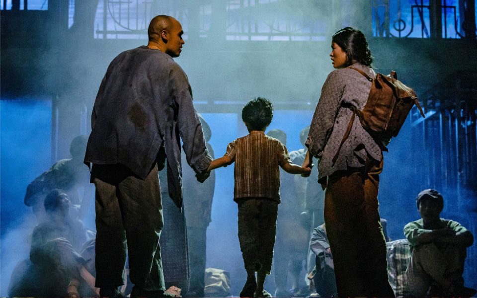 A man and woman in tattered dark clothes hold the hands of a small child. They face away towards people gathered and crouched on the floor.