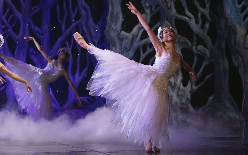 Sarah Chun in a white floaty dress and tiara with her arm and leg outstretched in a ballet pose. Around her is smoke and a winter woodland scene.