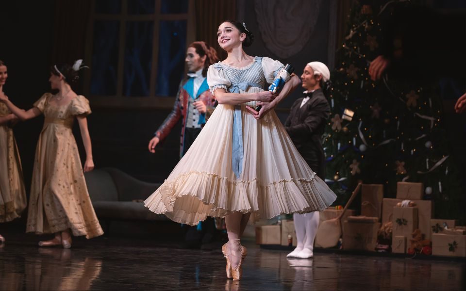 Rachael Gillespie as Clara in The Nutcracker wearing a white floaty dress with blue ribbon and holding a nutcracker cradled in her arms.