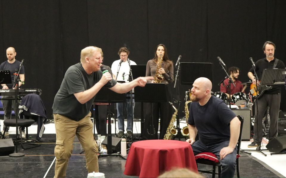 Simon Nock sings into a microphone as he wags his finger at Kenny Davies, who is sat at a small round table. The band plays behind them.