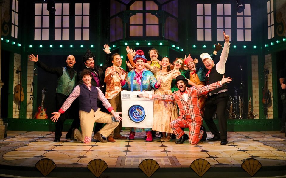 The full cast of Aladdin: The Rock 'n' Roll Panto smile, cheer and pose together on-stage, in front of a green building backdrop.