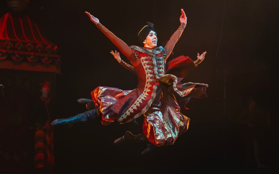 Albert González Orts in a gold-embellished red costume and black hat leaping into the air with his arms outstretched and his cloak billowing around him.