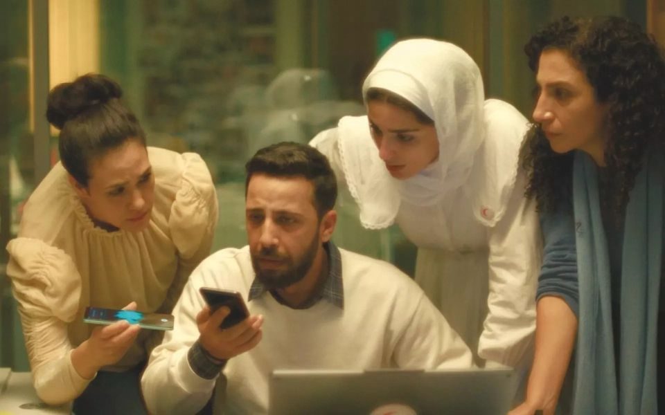 A man nervously speaks into a phone, as three other characters lean in closely, also holding a phone recording device.