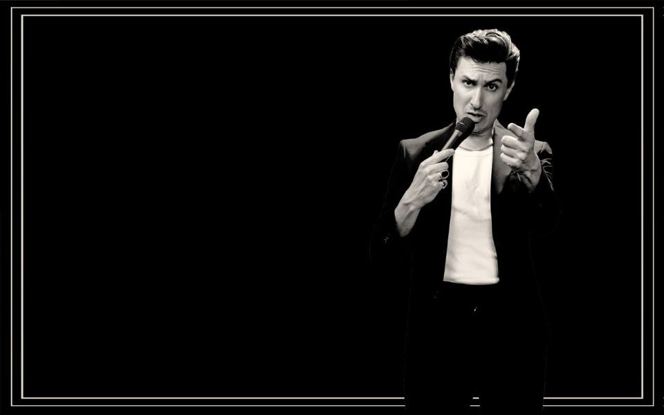 Tom Rosenthal with coiffured hair talks into a microphone, in black and white.