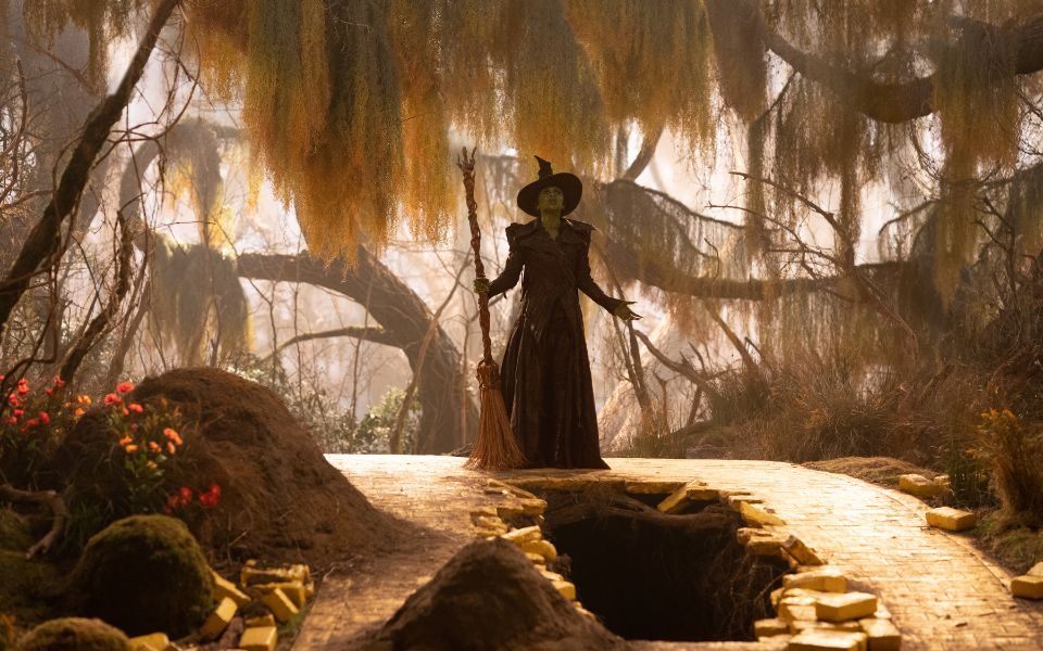 Cynthia Erivo as Elphaba stands above a hole in the yellow brick road, in a scene surrounded by weeping willow trees.