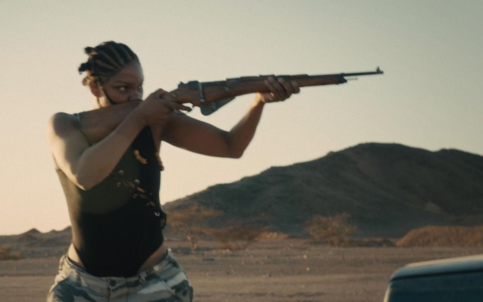 A woman holds up a rifle in a hazy desert setting.