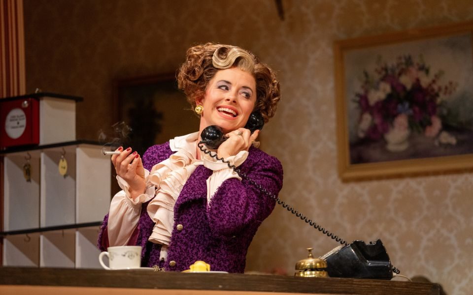 Sybil in Fawlty Towers answering the phone and smoking a cigarette in the lobby, smiling