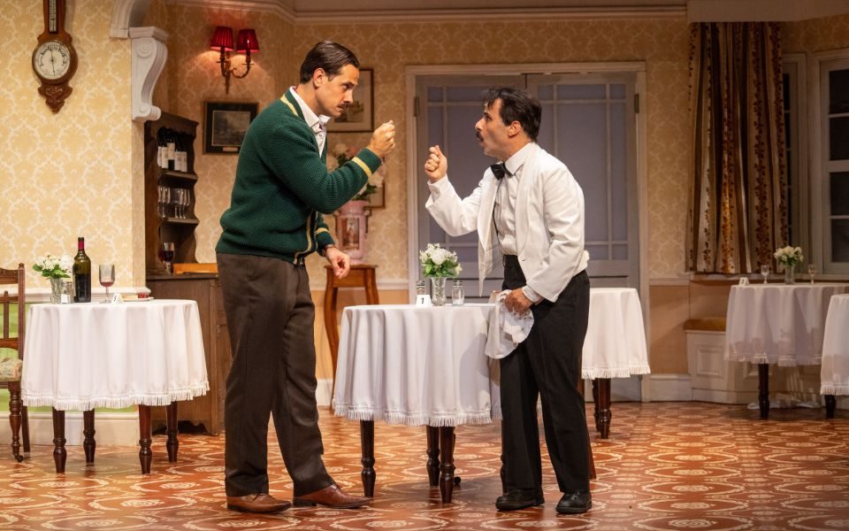 Danny Bayne as Basil in the iconic green cardigan talking passionately to Hemi Yeroham as Manuel in a white waiter suit and bow tie.