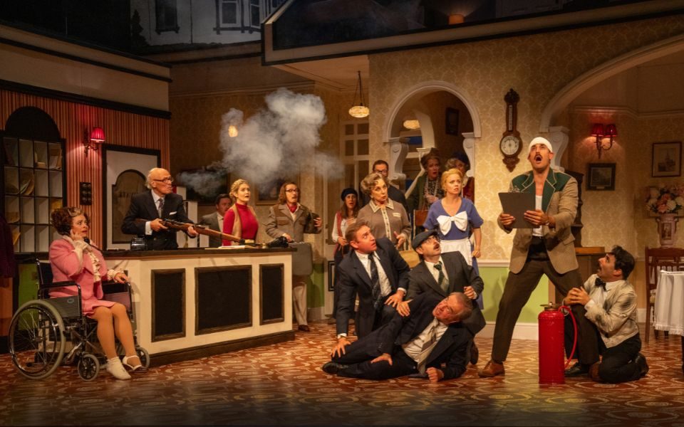 The cast of Fawlty Towers in the reception set. Someone holds a rifle that has gone off with a blast of smoke, some people are crouched on the floor, and everyone looks shocked. Manuel clutches Basil's leg as he shouts and holds a clipboard.