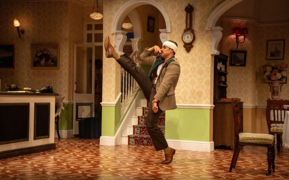 Danny Bayne as Basil Fawlty, wearing a suit and bandage on his head while kicking his leg high in the air to take a step.