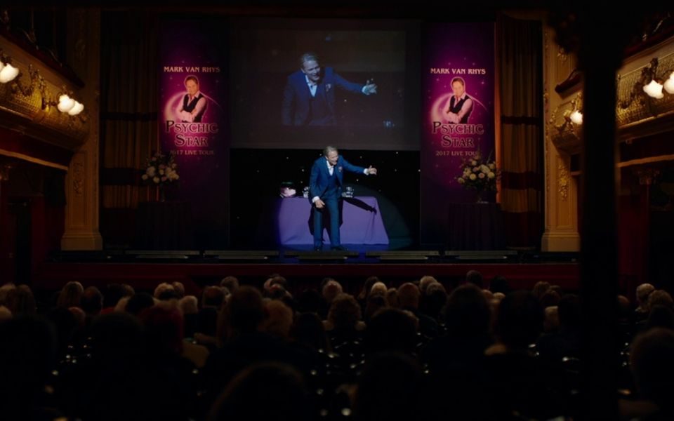 A man on stage under a spotlight, flanked by two purple posters reading 'Psychic Star, 2017 live tour' performs to a packed auditorium.