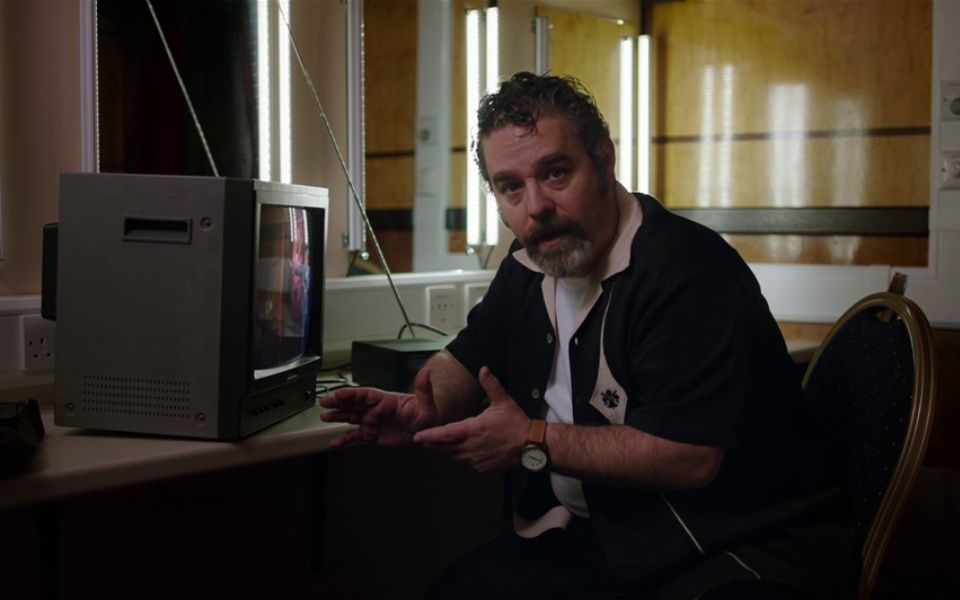 A man sits in a dim dressing room alongside a boxy television set.