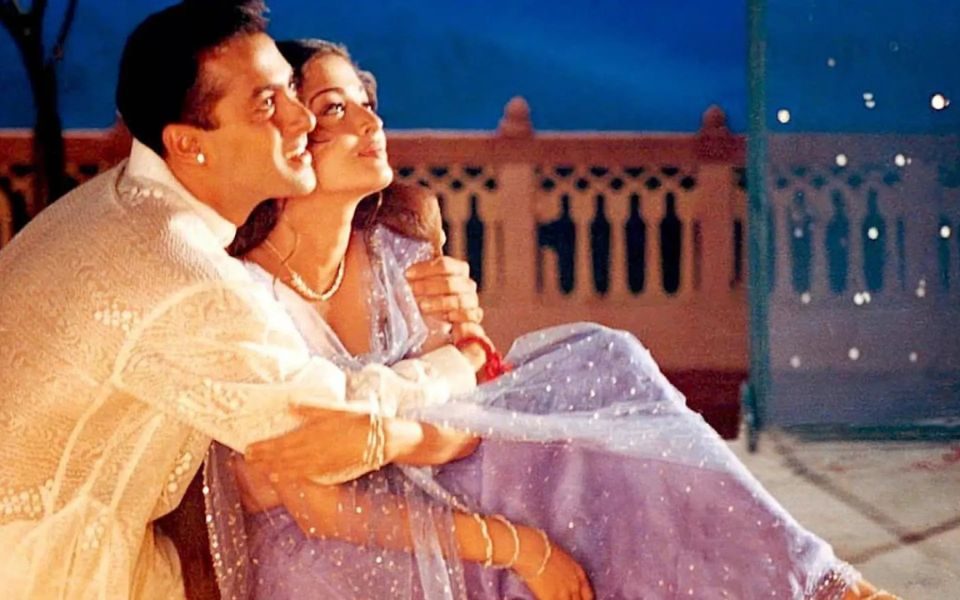 A couple in shimmering robes hold each other lovingly on a balcony.