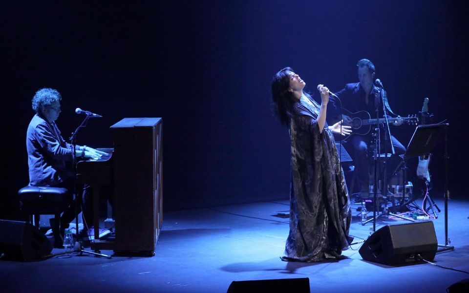 In moody blue lighting, Imelda May passionately sings on stage in a flowy dress, in front of an acoustic guitarist and beside a pianist.