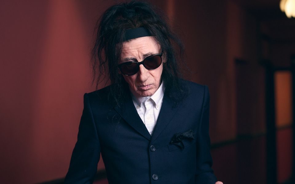 Dr John Cooper Clarke casually poses in a black suit, wearing his iconic sunglasses.