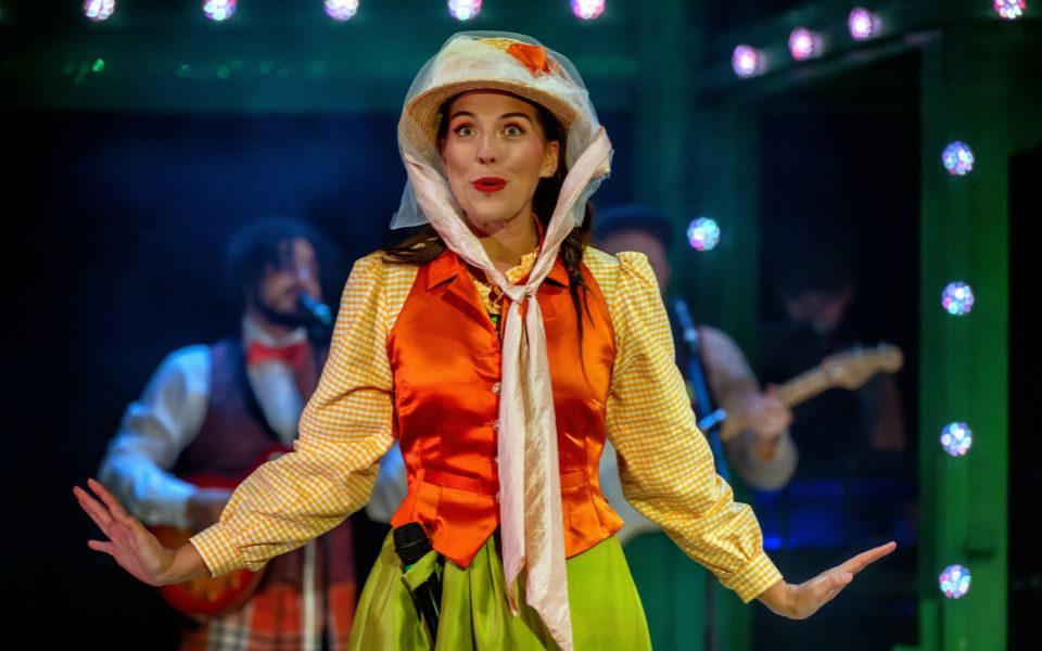 Laura Andresen Guimarães performs as Princess Peachblossom in a bonnet onstage, speaking to the crowd in Aladdin: The Rock n Roll Panto.