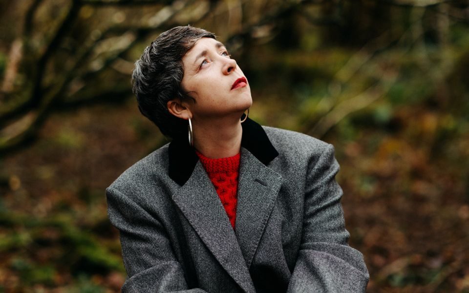 Lisa O'Neill sits in an autumnal forest, wearing a red jumper and grey blazer as she looks skywards.