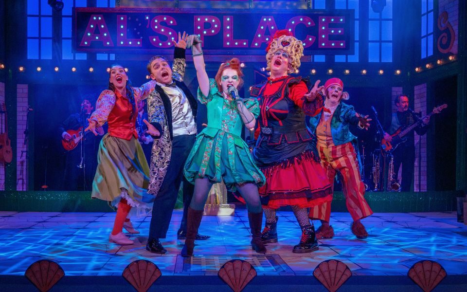 Lucy Wells as Genie singing in Aladdin: The Rock n Roll Panto, surrounded by singing cast members with outstretched arms.