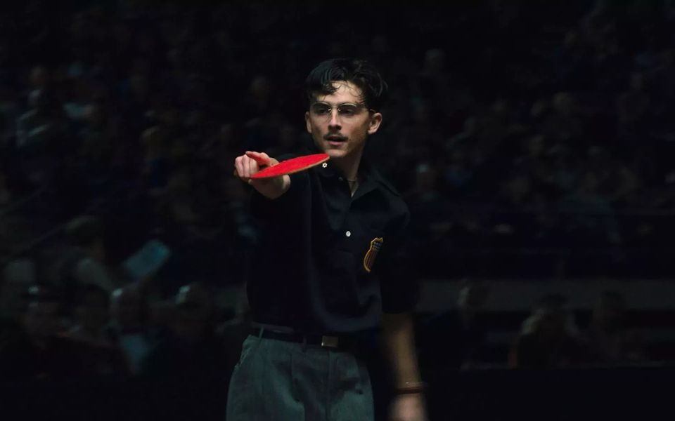 Timothee Chalamet wearing glasses as he plays table tennis in front of a low-lit crowd.