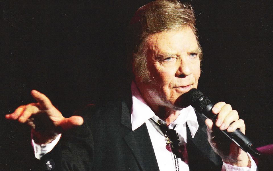 Marty Wilde sings into a microphone on stage, wearing a suit and bolo tie.