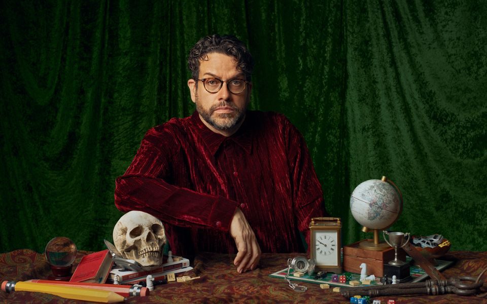 In the style of a Renaissance painting, Michael Spicer sits at a desk with his arm resting on a skull, as he wears a velvety red outfit in front of a dark green curtain. The desk also features pencils, books, dice, keys, a stopwatch and a globe.
