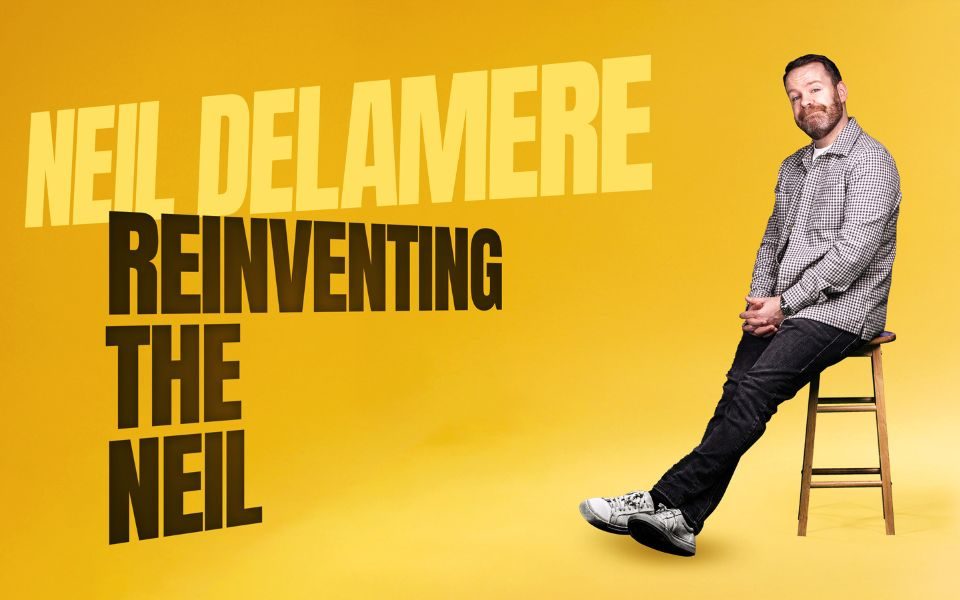 Neil Delamere perches on a high wooden stool in front of a yellow background. Text reads: Neil Delamere. Reinventing the Neil.