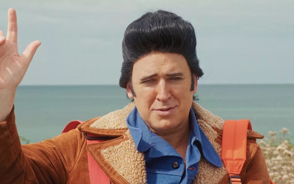 Tim Vine wears a black coiffured wig and furrows his brow at the camera.