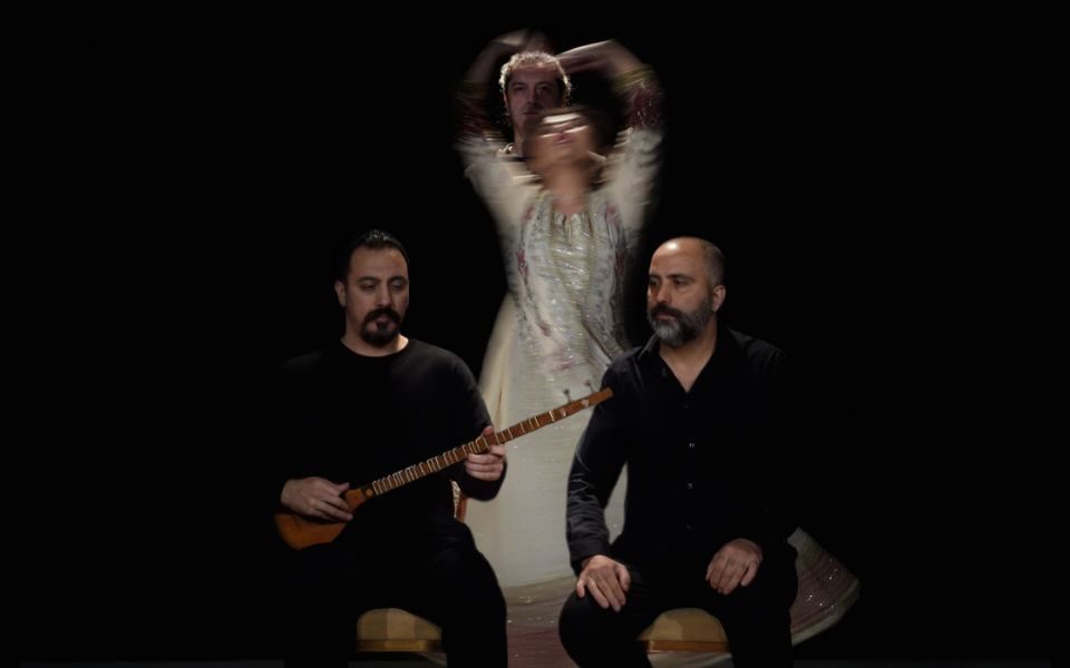 In a dark-lit room, two musicians sit on stools in front of a blurred dancer, in motion.