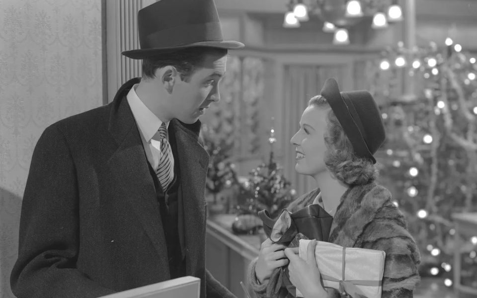 Black and white film still, featuring two characters in winter overcoats having a conversation, with lit Christmas trees and decorations in the background.