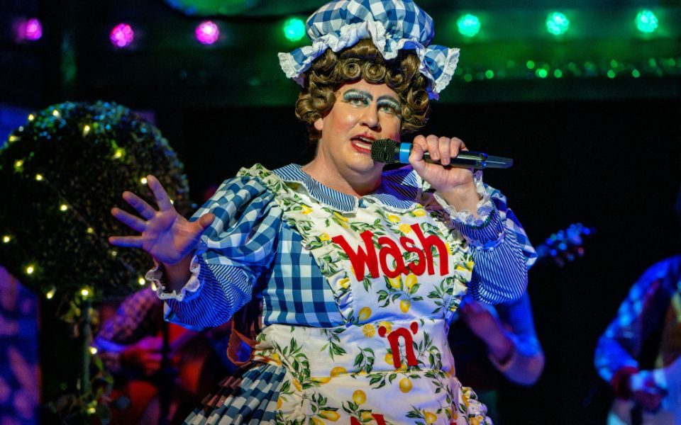 Simon Nock as the Dame in Aladdin: The Rock n Roll Panto, singing into a microphone in a dress and apron.