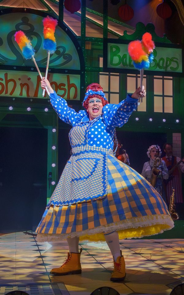 Simon Nock as the Dame on stage at City Varieties in Aladdin: The Rock n Roll Panto, holding colourful feather dusters and a wide dress.
