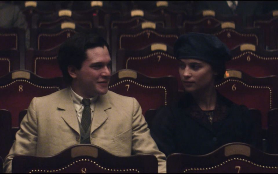 Kit Harington and Alicia Vikander in period costumes look at each other, sat in the auditorium of the City Varieties Music Hall.